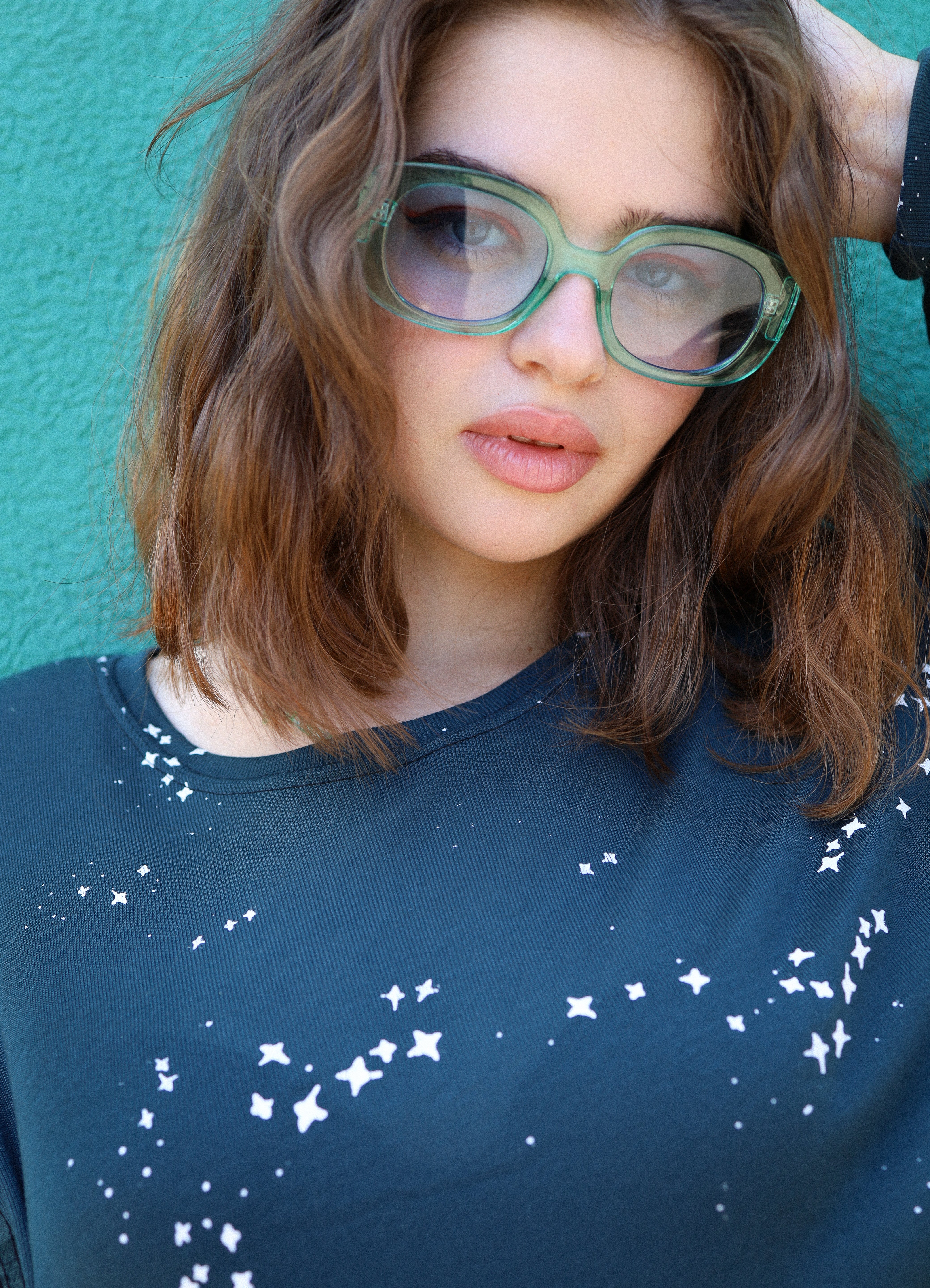 Person wearing green glasses and a blue shirt with white patterns against a teal background