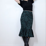 Woman wearing a black top and green patterned skirt against a white background