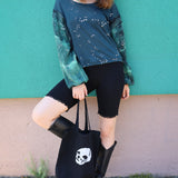 Woman holding a black tote bag with a white design against a teal wall.