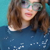 Person wearing green glasses and a blue shirt with white patterns against a teal background