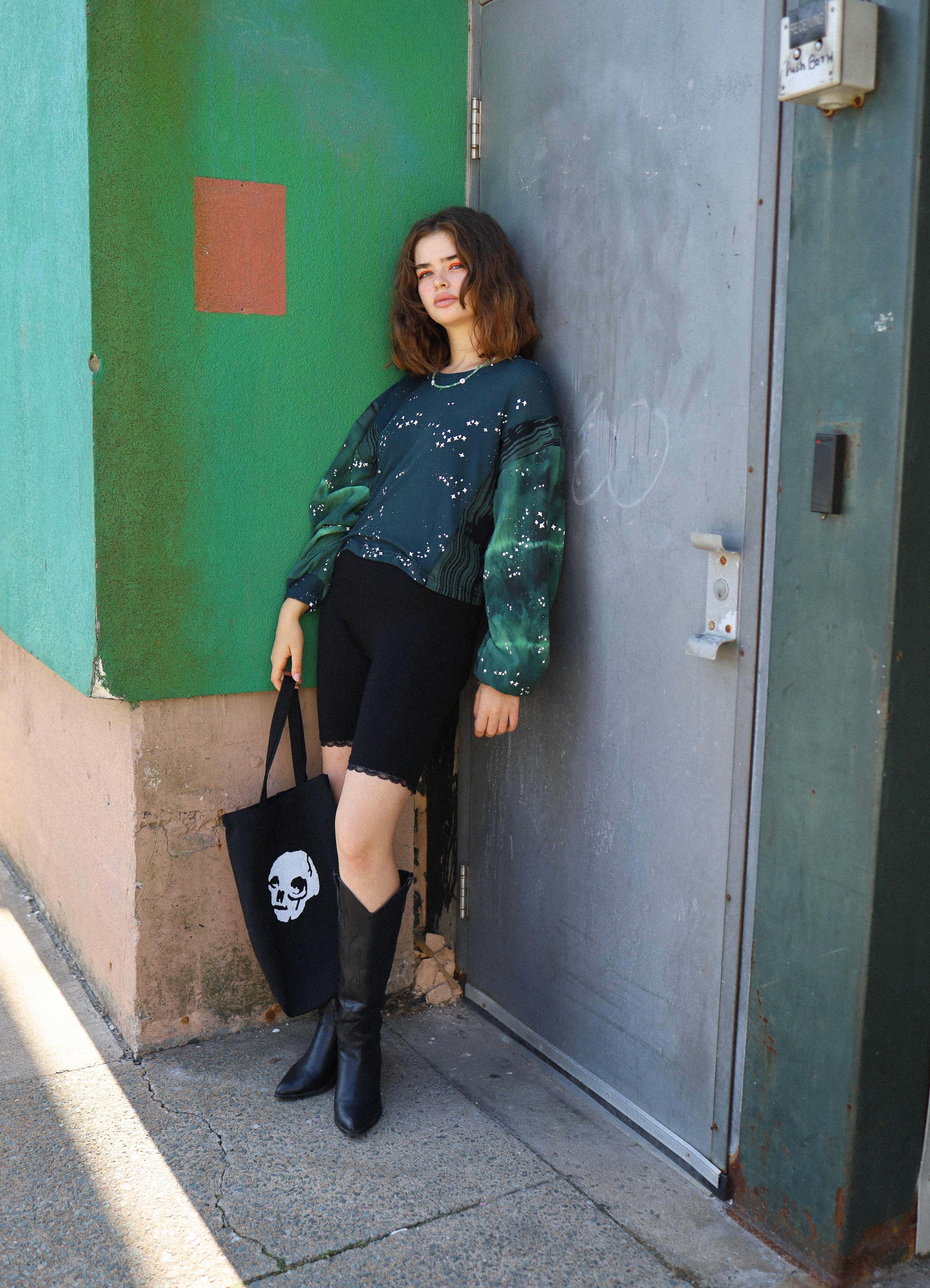 Woman standing against a green wall with a black door, holding a black bag with a white design.